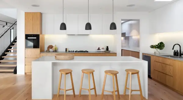 Moderna cocina blanca y madera clara. Isla central con taburetes, electrodomésticos integrados y suelo de madera.