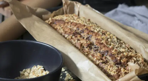 Frisch gebackenes Brot ohne Mehl, bedeckt mit gehackten Nüssen und Schokoladenstückchen, liegt in Backpapier. Daneben eine Schüssel mit Nüssen.