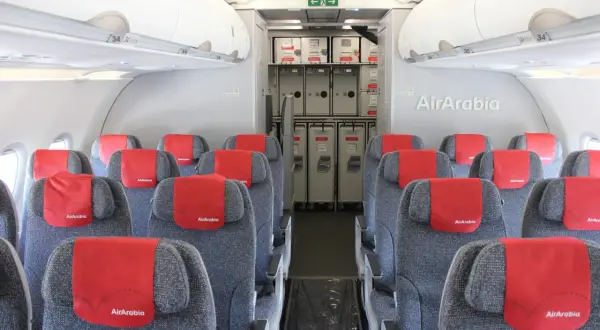 Wnętrze samolotu Air Arabia z rzędami szarych siedzeń i czerwonymi zagłówkami. W tle widać wózki pokładowe.