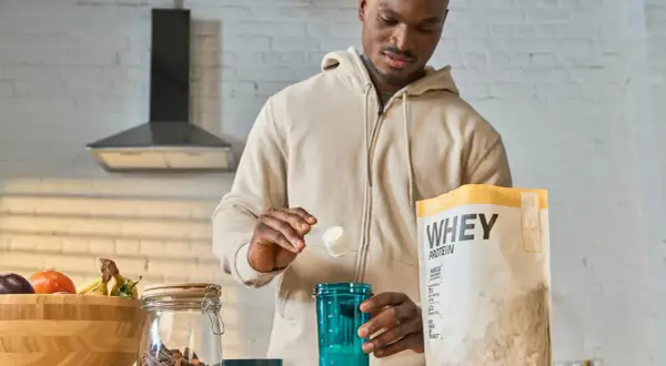 Mężczyzna przygotowuje odżywkę białkową po treningu. Na blacie stoi opakowanie WHEY PROTEIN, shaker i miska z owocami.