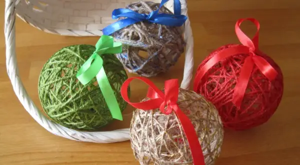 Palline decorative fatte con spago e colla vinilica, decorate con nastri colorati, appoggiate vicino a un cestino bianco.