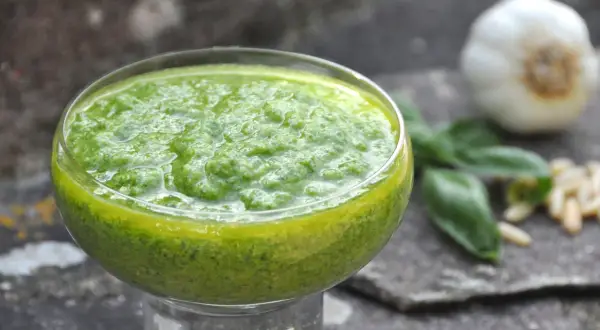 Pesto di basilico bimby in una coppetta, con aglio, pinoli e foglie di basilico sullo sfondo.