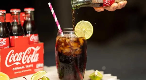 Coca-Cola nalewana do szklanki z lodem i limonką, obok wiśni i cytryn.
