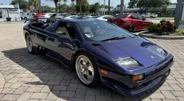 Fioletowy Lamborghini Diablo z jasnym wnętrzem, zaparkowany na brukowanym podjeździe, z palmami w tle.