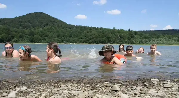 Dzieci kąpią się w górskim jeziorze, ciesząc się letnim dniem. Woda jest czysta, a wokół rozciąga się piękny, zielony las.