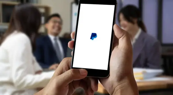 Dłoń trzyma smartfon z logo PayPal na białym ekranie, w tle rozmyte osoby w biurze.