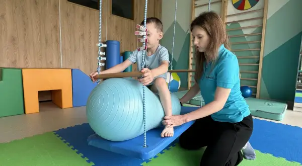Terapeuta pomaga dziecku na huśtawce z piłką terapeutyczną w sali integracji sensorycznej.