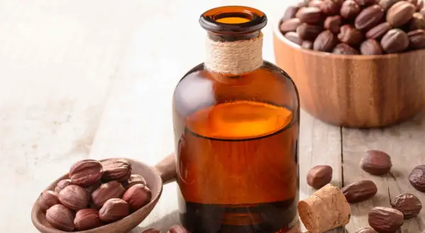 Butelka z bursztynowym płynem, obok drewniana łyżka z nasionami jojoby i miska pełna nasion. Naturalny olej jojoba na włosy.