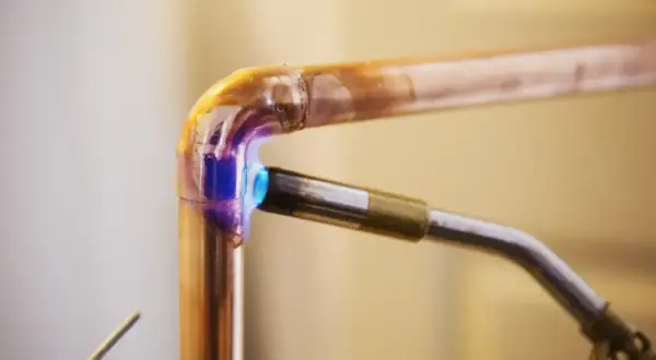 Un chalumeau chauffe un raccord coudé en cuivre pour le souder. La flamme bleue et violette illumine le métal.