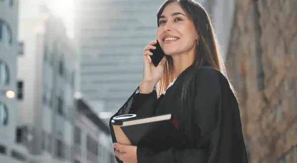 Uśmiechnięta prawniczka w todze rozmawia przez telefon, trzymając książkę i kawę.