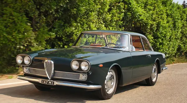 En mörkgrön Alfa Romeo Giulia Sprint GT kör längs en landsväg, en klassisk skönhet bland Alfa Romeo modeller.