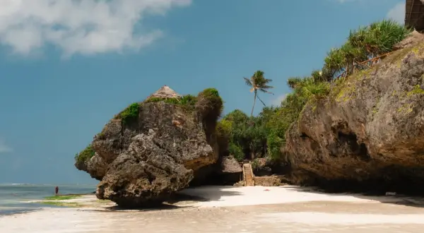 Zanzibar pora deszczowa. Skalista plaża z chatkami na klifie i palmą. W oddali widać samotną postać w czerwonej szacie.