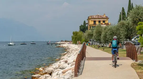 Ciclista sul lungolago, con barche e montagne sullo sfondo. Un assaggio di cosa vedere sul Monte Baldo, tra natura e relax.