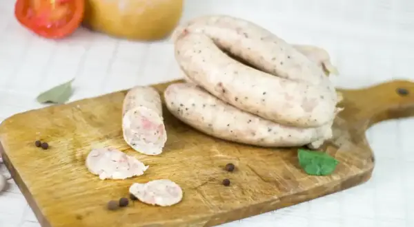 Biała kiełbasa na desce, gotowa do przygotowania według przepisu na 5 kg. W tle pomidor i cebula.
