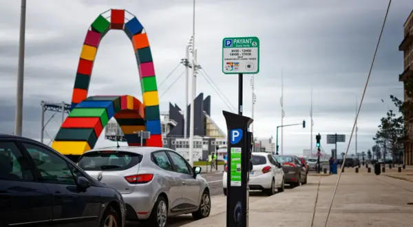 Panneaux de stationnement payant et horodateur, rappelant qu'une amende stationnement non payé est possible. Au loin, une arche colorée.