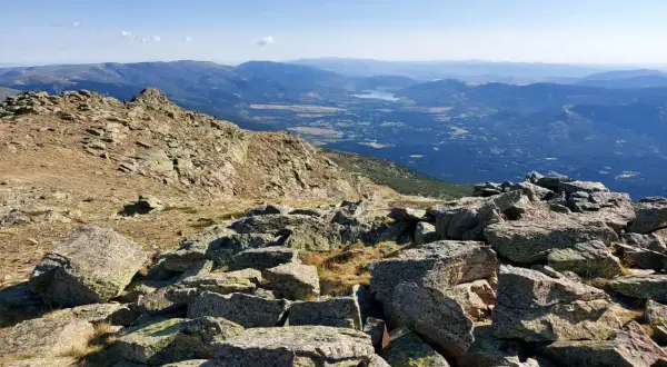 Vista panorámica de los picos de la Sierra de Guadarrama, con rocas cubiertas de líquenes en primer plano y un valle con un lago al fondo.