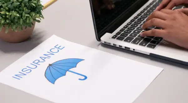 Osoba wpisuje dane na laptopie, obok leży dokument z napisem "INSURANCE" i symbolem parasola, sugerujący ubezpieczenie domu od wszystkich ryzyk.