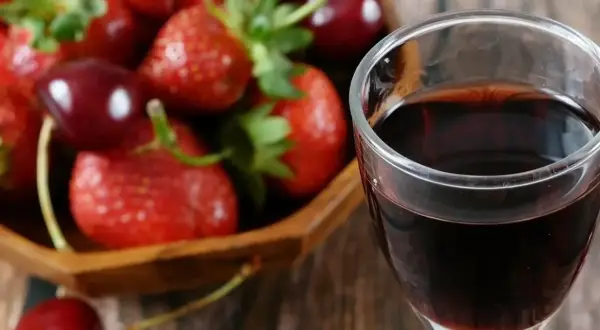 Szklanka ciemnego płynu, prawdopodobnie ratafia wódka, obok miski pełnej świeżych truskawek i wiśni.