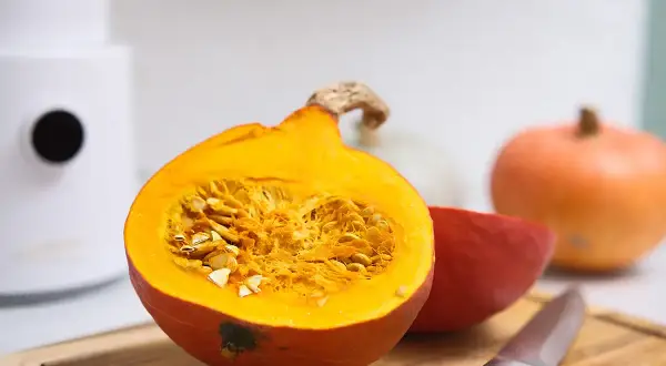 Pomarańczowa dynia przekrojona na pół, pełna nasion, obok inne odmiany dyni i nóż na desce.