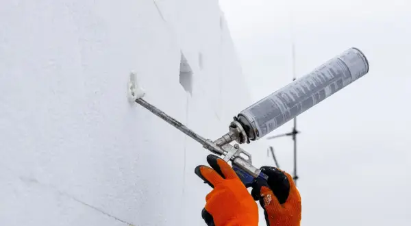 Ręce w pomarańczowych rękawicach używają pistoletu z pianką do wypełnienia otworu w białej płycie izolacyjnej. To jak tworzenie tulei dystansowej.