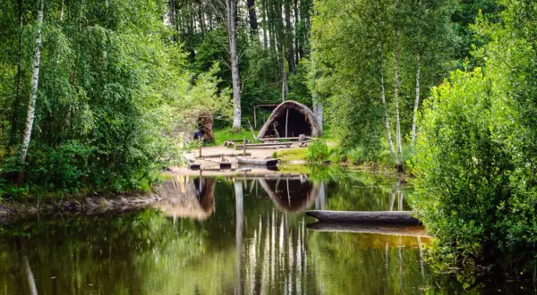 En stilla sjö speglar grönskande träd och en hydda vid Årås kvarn. En stockbåt vilar vid strandkanten.
