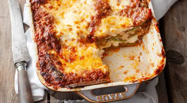 Lasagne alla bolognese appena sfornate, dorate e filanti, in una teglia Staub. Un coltello vintage è appoggiato accanto.