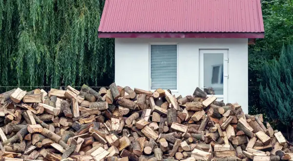 Duża sterta drewna opałowego przed białym budynkiem, z zielonym lasem w tle. Odległość budynku od lasu jest niewielka.