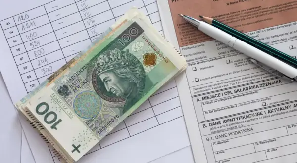 Stos banknotów 100 zł leży na formularzu PIT. Obok arkusz z danymi i ołówki.