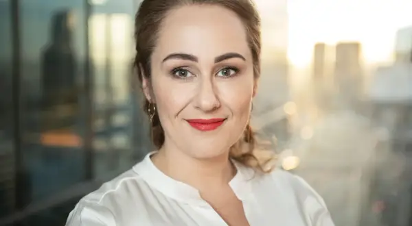 Portret amerykański kobiety z rudymi włosami, w białej bluzce, z czerwonymi ustami, na tle miasta.