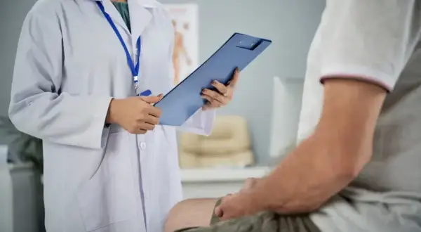 Lekarz w białym kitlu trzyma teczkę, rozmawiając z pacjentem. To wizyta, podczas której lekarz bada prostatę.