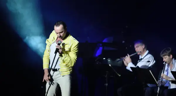 Wokalista w żółtej kurtce śpiewa na scenie, podczas gdy muzycy grają na instrumentach dętych. To koncert queen symfonicznie.
