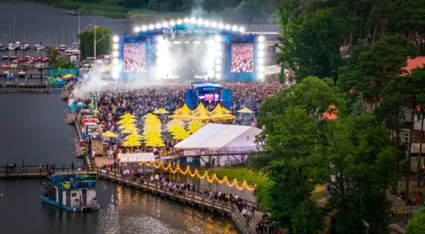 Tłumy ludzi na festiwalu Rap Stacja, scena z ekranami i dymem, żółte parasole, łódź na wodzie.