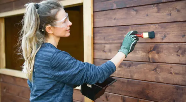 Donna con occhiali protettivi applica una vernice scura su una parete di legno, un tipo di pittura che protegge e abbellisce.
