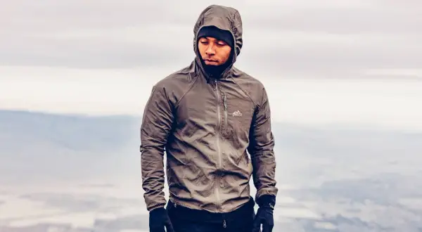 Mężczyzna w brązowej kurtce softshell z kapturem i czarnej czapce, na tle mglistego krajobrazu.