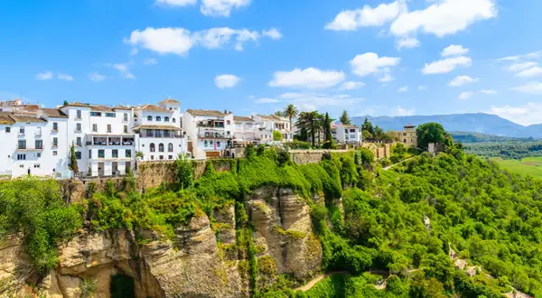 Vita hus i Ronda, Spanien, klättrar uppför en grönskande klippa under en blå himmel med moln.