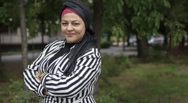 Zeina Mourtada, med en svart och röd turban och en randig jacka, står med armarna i kors utomhus.
