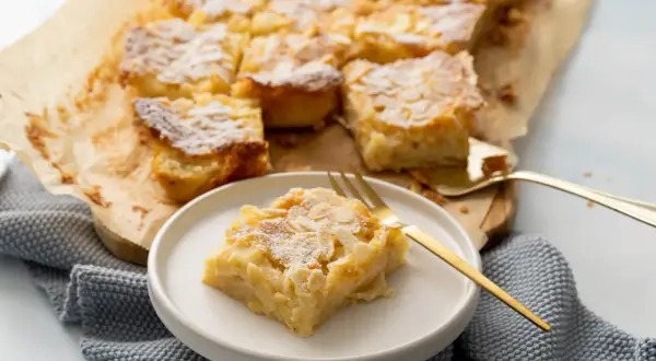 Ein saftiger Apfelkuchen vom Blech, bestreut mit Puderzucker und Mandelblättchen. Ein Stück liegt auf einem Teller, bereit zum Genuss.