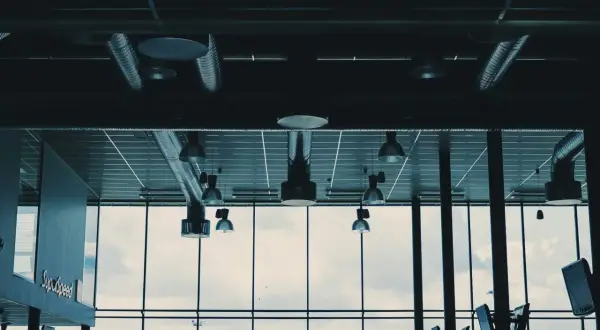 Synliga ventilationsrör och belysning i taket, med stora fönster som visar en molnig himmel och en grönskande kulle.