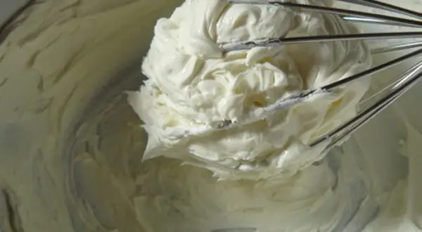Un fouet de cuisine retient une généreuse portion de glacage mascarpone crémeux, prêt à garnir un gâteau.