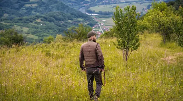 Uomo con fucile attraversa prati fioriti, godendosi la vista dei passi di montagna e della valle sottostante.