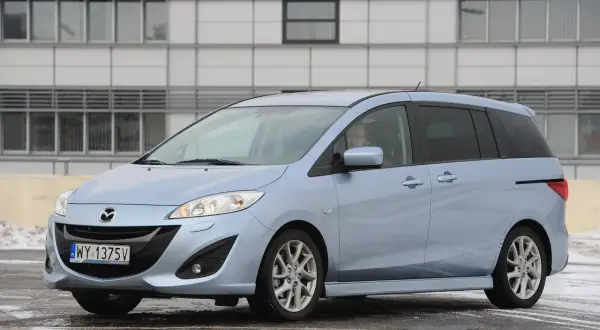 Jasnoniebieska Mazda 5 z nowymi żarówkami w reflektorach, zaparkowana przed nowoczesnym budynkiem.