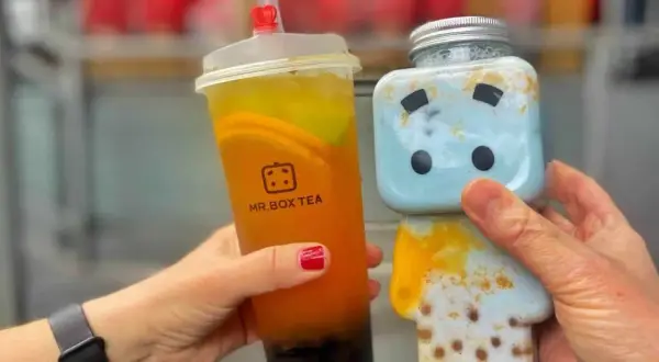 Zwei Hände halten leckere Getränke: ein Bubble Tea von Mr. Box Tea in Berlin und ein süßer blauer Drink in einer Figurenflasche.