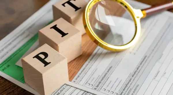 Drewniane klocki z literami "PIT" leżą na formularzu podatkowym. Złota lupa przybliża szczegóły. Czas zacząć myśleć, od kiedy można rozliczać PIT.