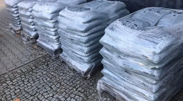 Stosy worków z asfaltem na paletach, owinięte folią, na kamiennym podłożu.