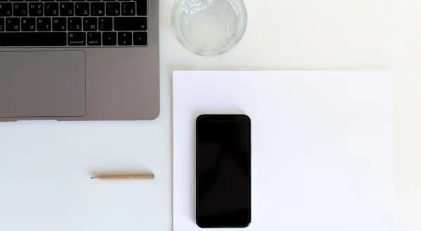 Przygotowanie do naklejania szkła na telefon. Obok laptopa i szklanki wody leży czarny smartfon na białej kartce.