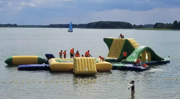 Rodzinna zabawa na wodnym placu zabaw na Mazurach. Dzieci w kamizelkach ratunkowych wspinają się i ślizgają po dmuchanych konstrukcjach.
