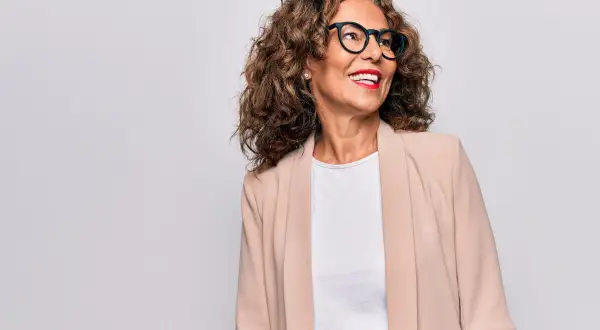 Donna sorridente con occhiali e blazer beige, un esempio di come vestirsi a 50 anni con stile.