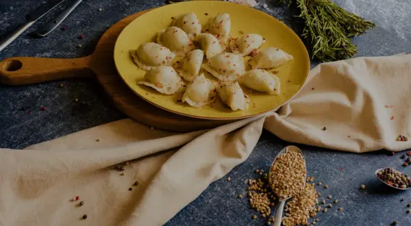 Talerz żółtych pierogów ruskich, posypanych papryką. Obok łyżka kaszy gryczanej i przyprawy. Zastanawiasz się nad pierogi ruskie kcal?