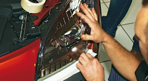 Ręce zabezpieczają reflektor Mazdy 3 taśmą przed wymianą żarówki H7.