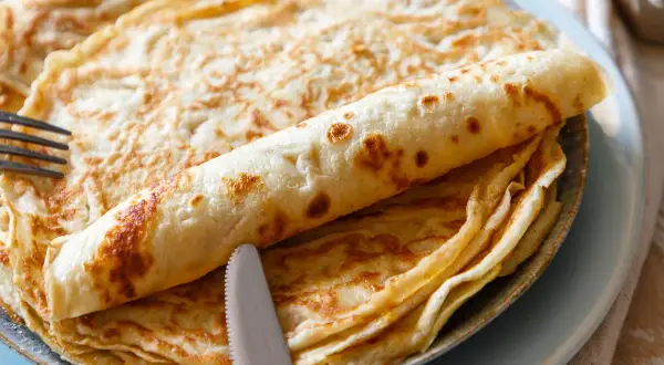 Ein Stapel goldbrauner Eierkuchen, einer davon aufgerollt, liegt auf einem Teller. Ein Messer und eine Gabel liegen daneben. Perfekt für ein einfaches Eierkuchen Rezept.